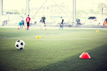 Soccer ball on green artificial turf with blurry kid soccer team training. Blurry kid soccer player jogging between marker cones and control ball with soccer equipment in football academy.