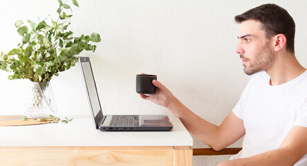 Retrato de un hombre adulto y apuesto caucásico sentado frente a su laptop y bebiendo una taza de café mientras la usa en un apartamento brillante y blanco con luz natural.