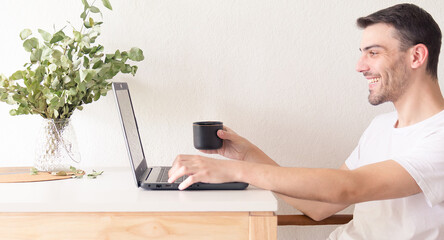 Retrato de un hombre adulto y apuesto cauc&aacute;sico sentado frente a su laptop y bebiendo una taza de caf&eacute; mientras la usa en un apartamento brillante y blanco con luz natural.