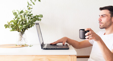 Retrato de un hombre adulto y apuesto caucásico sentado frente a su laptop y bebiendo una taza de café mientras la usa en un apartamento brillante y blanco con luz natural.
