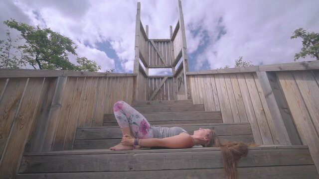 Side view of young athletic girl doing yoga on wooden stairs garden, static