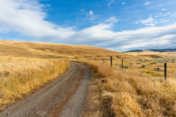 Country, dirt road between fields with yellow grass, near a fence with barbed wire to the hill