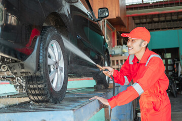 Fototapeta na wymiar Smiling Asian male car cleaner wearing red uniform and hat is spraying water on a car in the car salon