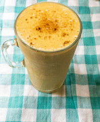 Above view of a pumpkin spice smoothie in a clear glass mug on a green and white checkered tablecloth. Cinnamon is sprinkled on tope of the smoothie.