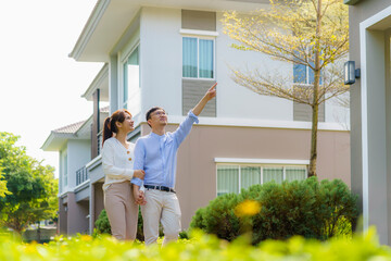 Portrait of Asian couple walking hugging and pointing together looking happy in front of their new...