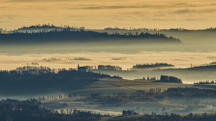 Sun light rays during sunshine peaking through morning mist on a wavy landscape with a church on top of a hill, Jeseniky, Czech Republic.