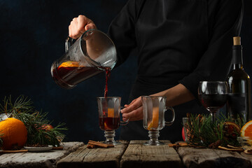 Chef pours aromatic mulled wine with fragrant spices into glasses on rustic table with festive composition background. Concept of preparing hot winter alcoholic drink. Frozen motion. Classic recipe
