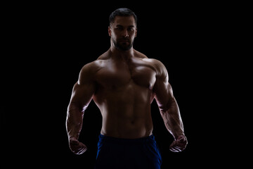 Fototapeta na wymiar Portrait of a bodybuilder standing isolated on black background in a shadow with clenched fists to show off his muscles