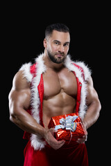 Muscular sexy sportsman in a red Santa Claus costume over naked body is holding a Christmas gift isolated on black background