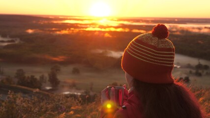 woman Traveler holds a metal mug with hot coffee in her hands and looks at sunrise from mountain. free girl hiker admiring landscape. Tourist is drinking tea from a mug in sun. travel, camping