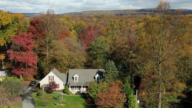Cape Cod home in colorful woods. Descending aerial drone shot of red, yellow, orange fall foliage in forest in United States, USA. House establishing shot.