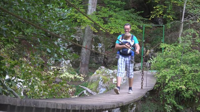 Father with holding child, baby wearing, passing on suspended wooden bridge, dangerous bridge above river mountain