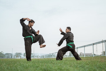 Two fighters in pencak silat uniform pose with stances in an outdoor background