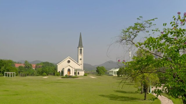 Catholic style church building without a cross surrounded with green grass fields on a bright clear day.. Aerial dolly left from behind a green tree.