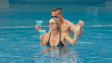 The loving couple hugs and kisses, drinking blue cocktail alcohol liquor in swimming pool at hotel outdoor. Portrait of caucasian man and woman.