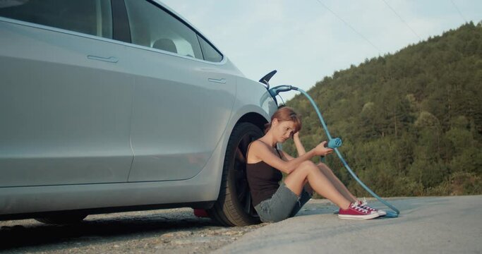 Sad girl sitting on the ground next to her empty electric car. Holding charging cable with nowhere to plug.