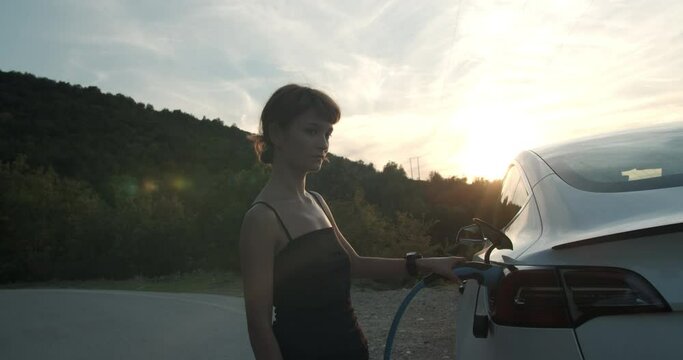 Girl holding the charging cable in her electric car during sunset. Handheld video.