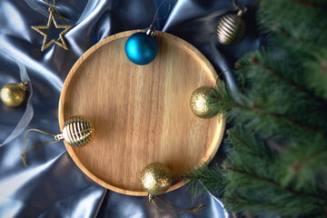 Top view of empty wooden plate decorate with gold and blue baubles on blue silver satin silk cloth with Christmas tree as foreground. Festive decoration for New year’s day and Merry Christmas season