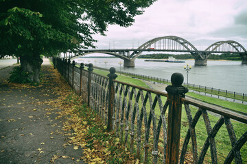 An old embankment with large old linden trees and cast-iron railings overlooking the road bridge over the river, under which a barge floats against the background of an autumn cloudy sky.Rybinsk city.