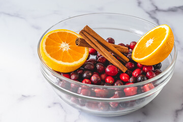 Fresh cranberries, cinnamon and oranges close up in a glass bowl on marble background. Ingredients for cooking cranberry sauce