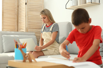 Young woman working on laptop while son drawing in living room. Home office concept