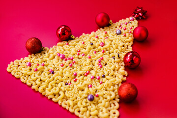 Christmas tree made of raw italian pasta with red balls isolated on red background. Winter holidays concept