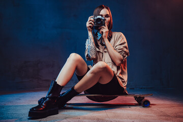 Sportive and stylish girl dressed in casual clothing with short haircut and tattoo sits on skateboard with camera in studio room.