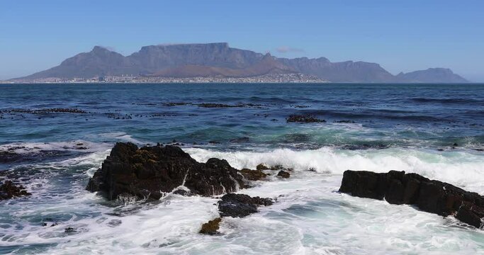 Waves crashing on rock on the shore of Robben Island with Table Mountain and Cape Town in the backdrop. 