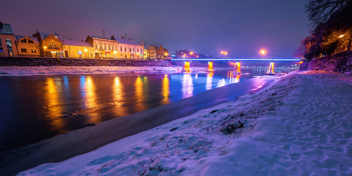 uzhhorod, ukraine - 26 DEC, 2016: old town on a christmas night. beautiful cityscape by the river. snow on the embankment. bridge and lanterns glowing in the distance