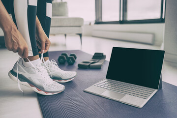 Home workout with laptop. Online fitness class . Woman getting ready for exercising tying her running shoes with technology device streaming on black screen.