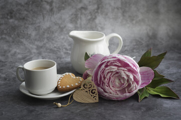 On a gray background: a pink peony flower, a wooden heart, a jug, a cup of coffee with milk and cookies. Congratulations on Valentine's Day, Happy Mother's Day.