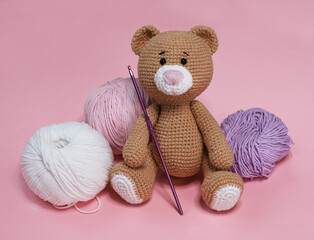 Hand knitted brown bear with a pink nose sitting among balls of thread on a pink background