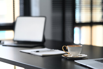 A white coffee cup is putting on contemporary workspace with empty screen computer laptop on blace wooden table.