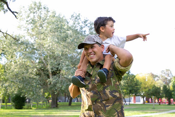 Exited little boy sitting on dad neck and pointing away. Caucasian father holding son legs,...