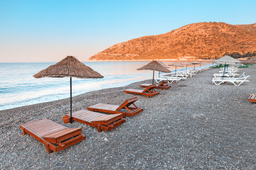 Sunbeds and sun umbrellas await vacationers on the shingle beach at Ovabuku beach on the Datca Peninsula in Turkey. The photo was taken in the early morning at sunrise