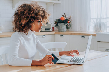 Dark skinned lady with kinky hair in white typing on keyboard holding blue credit card enjoys shopping online in kitchen closeup