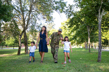 Obraz premium Front view of happy family walking together on meadow in park. Father wearing military uniform and showing something to daughter. Long-haired mom smiling. Family reunion and returning home concept