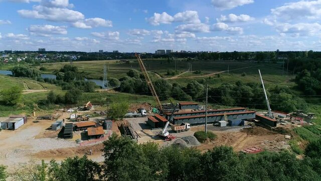 Aerial top down view of construction yard bridge building project in showing front construction crane, truck, piling machine over unpaved soil and the infrastructure ready to move.