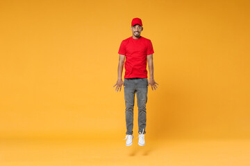 Fototapeta premium Full length fun delivery employee african man in red cap blank print t-shirt uniform workwear work courier dealer service on covid-19 virus concept jump run walk isolated on yellow background studio.