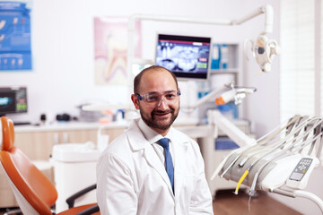 Confident dentist in stomatology cabinet with orange equiptment wearing dental uniform. Medical specialist in oral hygiene wearing lab coat looking at camera in dentistry office.