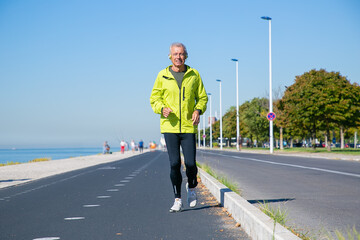Happy excited mature man in wireless headphones jogging along river bank outside. Senior jogger training for marathon. Front view. Activity and age concept
