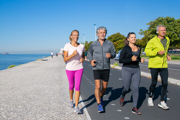 Happy mature people wearing sports clothes, jogging along river bank. Full length, copy space. Retirement or active lifestyle concept
