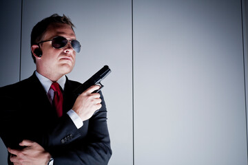 Dark portrait of caucasian man in dark suit with headset, sunglasses and a hand gun, bodyguard. Grey background with copy space. - obrazy, fototapety, plakaty