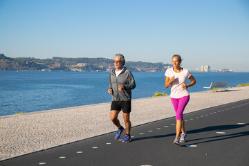 Positive mature couple keeping active lifestyle and jogging along river bank in morning. Retired people activity concept