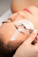 Closeup view of young caucasian woman having eyelash extension procedure in beauty salon. Beautician glues eyelashes with tweezers
