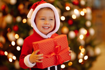 Little boy in santa costume with christmas gift. Happy child holding red present  box on the background of Christmas lights and tree. Winter holiday, New Year.