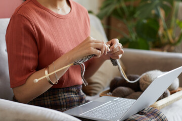 Close-up of woman sitting on sofa with laptop on her knees and knitting wool sweater at home