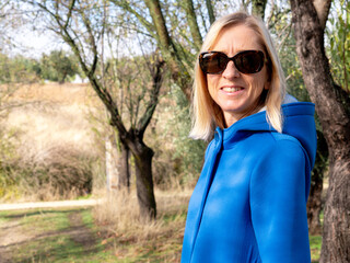 BLONDE DIE IN A BLUE COAT, JEANS, SNEAKERS AND SUNGLASSES, IN A FIELD OF OLIVE TREES IN AUTUMN, SPAIN, POSING HAPPILY NEXT TO A TREE