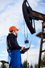 Oil man holding notebook and looking at oil pump rocker-machine. Male worker in uniform and helmet...