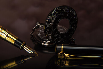 Fountain pen and pocket watch on mirrored surface with blurred background, selective focus.